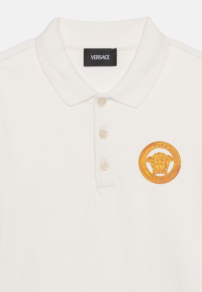 Λευκό πόλο πουκάμισο με τρία κουμπιά και χρυσό έμβλημα Μέδουσας στην αριστερή πλευρά του στήθους, με μαύρη ετικέτα της μάρκας Versace μέσα στον γιακά.