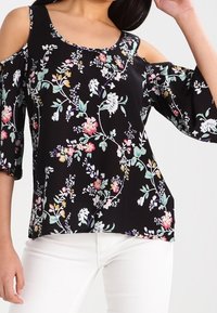 Blusa nera floreale con design cold-shoulder. Presenta vivaci motivi floreali in rosa, bianco e verde su un tessuto setoso. Maniche a tre quarti.