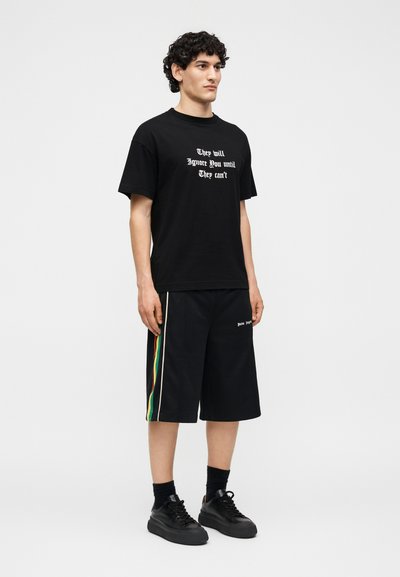 Άνδρας με σγουρά μαλλιά, που φοράει μαύρο T-shirt με λευκό κείμενο "They will Ignore You until They can't", μαύρες βερμούδες με πλευρικές ρίγες και μαύρα αθλητικά παπούτσια.