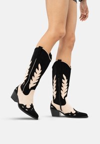 Piernas vestidas con botas vaqueras de ante negras y beiges hasta la mitad de la pantorrilla, con patrones en forma de hoja y tacones bajos, contra un fondo blanco.