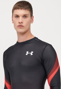 T-shirt de sport noir à manches longues avec des accents rouges et un logo Under Armour blanc sur la poitrine. Tissu lisse, design ajusté et col rond.