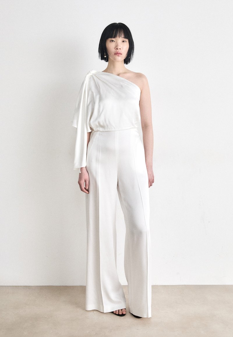 White one-shoulder jumpsuit with loose fit; satin texture; wide, flowing legs; features a draped sleeve accent.