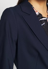 Námořnický blejzr s přizpůsobeným designem, s klopou typu notch a hladkou texturou látky; v kombinaci s černou blůzou s volány a květinovými akcenty.