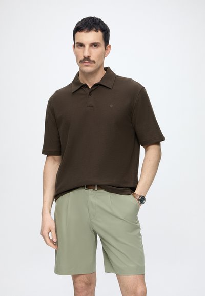 Homme aux cheveux foncés et moustachu portant un polo marron, un short vert clair, une ceinture marron et une montre-bracelet, debout avec une main dans la poche.