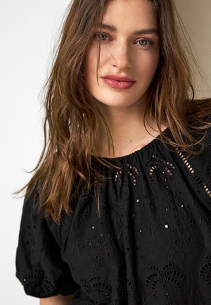 Tânără cu pielea deschisă la culoare, ochii verzi și părul lung și castaniu, purtând o bluză neagră brodată, privind ușor în lateral cu un zâmbet blând.