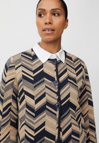 Giacca trapuntata con motivo a zigzag in blu navy e beige. Presenta un colletto bianco e bottoni blu navy lungo la parte anteriore. Tessuto testurizzato.