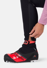 Schwarzer und roter Langlaufski-Schuh mit glänzender Oberfläche, ausgestattet mit einem Klettverschlussriemen und einem Textilkragen. Reißverschlussdetails im Knöchelbereich.