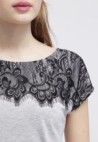 Šedé tričko s krátkými rukávy, které má černou krajkovou vrstvu na výstřihu s květinovými vzory a zvlněnými okraji.