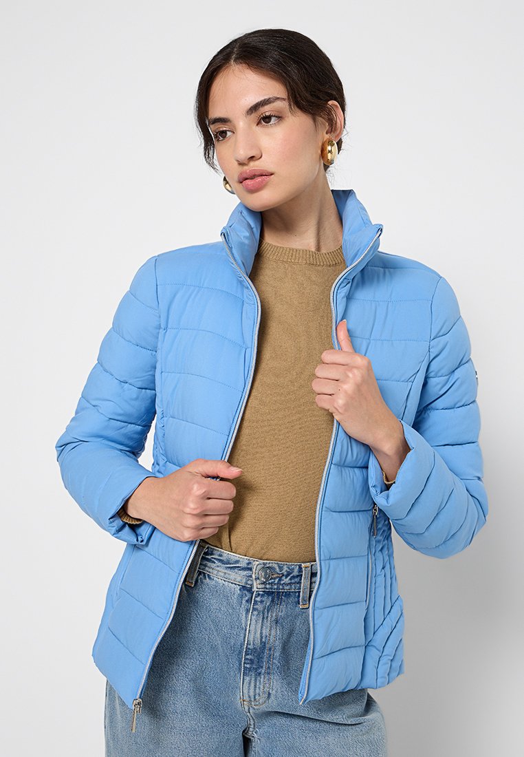 Hellblaue, gesteppte Jacke mit hohem Kragen, Reißverschluss und seitlichen Taschen, getragen über einem braunen Pullover und kombiniert mit Jeans.