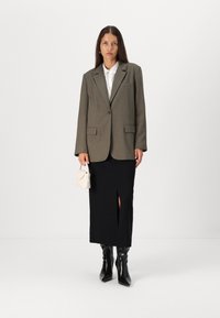 Blazer vert olive avec des revers pointus, jupe noire avec fente avant, bottes noires jusqu'aux genoux et un petit sac à main crème.