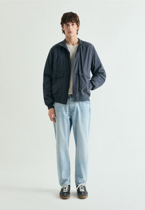 Jonge man staand, gekleed in een marineblauwe jas, wit T-shirt, lichtblauwe spijkerbroek en zwarte sneakers met witte veters, tegen een effen achtergrond.