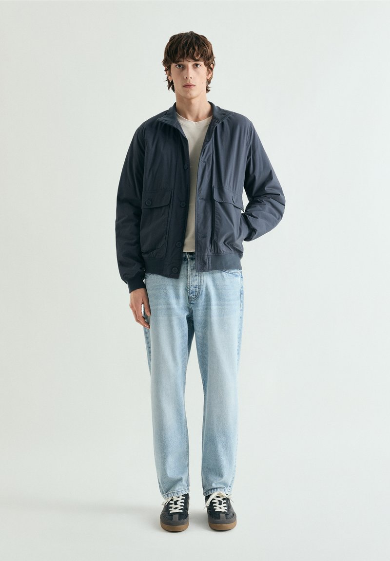 Ung mand stående iført en navyblå jakke, hvid T-shirt, lyseblå jeans og sorte sneakers med hvide snørebånd, foran en ensfarvet baggrund.