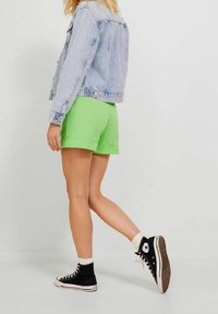 Giacca di jeans azzurro chiaro, pantaloni corti verde con risvolti, e sneakers nere alte con dettagli bianchi. Lo sfondo è bianco uniforme.