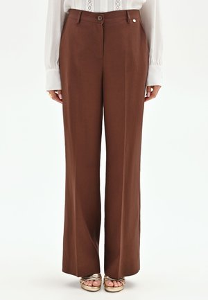 Pantalon marron taille haute à jambes larges avec fermeture par bouton et zip, porté avec un chemisier blanc à manches longues et des sandales dorées à bouts ouverts.