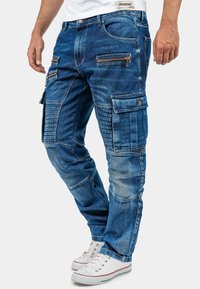 Pantalons cargo en denim bleu avec plusieurs poches zippées, panneaux de cuisse texturés et design à jambe fuselée, associés à des baskets blanches.
