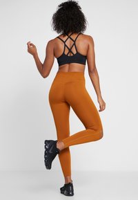 Reggiseno sportivo nero con spalline incrociate, abbinato a leggings aderenti, a vita alta, color ruggine e scarpe sportive nere. Tessuto liscio.