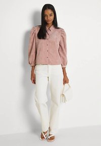 Γυναίκα φορώντας ένα ροζ πουά μπλουζάκι με φουσκωτά μανίκια και κουμπιά, λευκά παντελόνια ψηλής μέσης, λευκά σανδάλια και κρατώντας μια μικρή λευκή τσάντα.