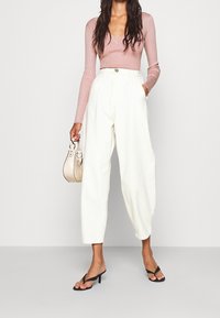 Lichtroze ribbeltop met lange mouwen, gecombineerd met loszittende witte broek; model houdt een kleine beige tas vast en draagt zwarte sandalen.