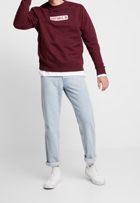 Vinfärgad sweatshirt med "JUST DO IT." tryckt i vitt, kombinerad med ljusblå jeans med uppvikta ärmar och vita sneakers. Enkel, avslappnad design.