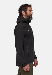 Veste noire imperméable avec capuche, dotée d'un zip sur le devant et de poches latérales. Le design est épuré avec des textures contrastantes et des coutures visibles.