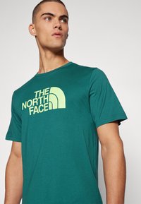 Teal lyhytnäppisen t-paidan, joka on valmistettu pehmeästä kankaasta ja jossa on iso, kontrastiväri vaalea keltainen logo, jossa lukee "The North Face."