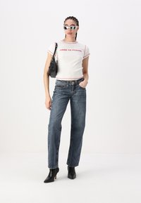 Baltas trumpas rankovės marškinėliai su raudonu apvadu ir užrašu "SUNKIAS ĮTIKTI", derinami su aukštu lygmeniu mėlynais džinsais ir juodais aulinukais.