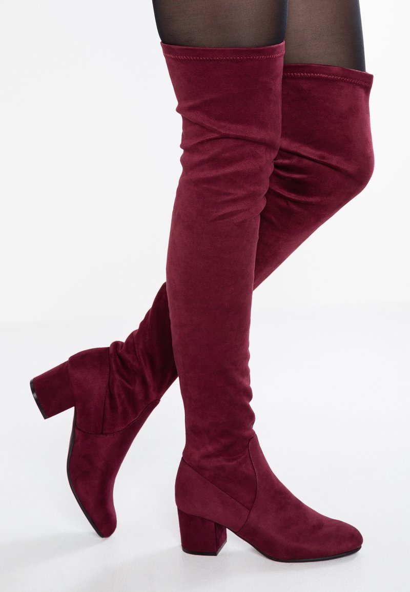 Femme portant des cuissardes en daim bordeaux à talons blocs avec des collants noirs transparents sur fond blanc.