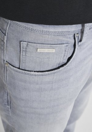 Bolsillo de jeans de mezclilla azul claro con una pequeña etiqueta metálica que dice "ANTONY MORATO" en una persona que lleva una camiseta negra.