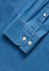 Chemise en denim bleu clair à manches, ornée de deux boutons blancs, d'un poignet replié et d'un détail de couture visible. Texture lisse avec un éclat subtil.