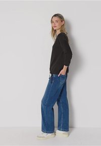 Musta pitkähihaine paita, jossa on sivukiristys, yhdistettynä vaalensinisiin leveälahkeisiin farkkuihin ja valkoisiin tennareihin, yksinkertaisella taustalla.