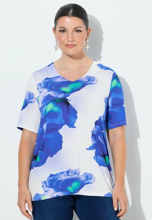 Femme aux cheveux foncés portant un haut blanc à col en V orné de grands motifs floraux bleus et verts, associé à un pantalon bleu foncé.