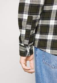 Zelená a černá kostkovaná flanelová košile s bílými akcenty, se zapínacími manžetami a volným střihem, v kombinaci se světle modrými džíny.