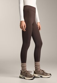 Bruine hooggetailleerde leggings van rekbaar materiaal, met een zijzak, gecombineerd met chunky bruine sneakers en beige gebreide sokken.