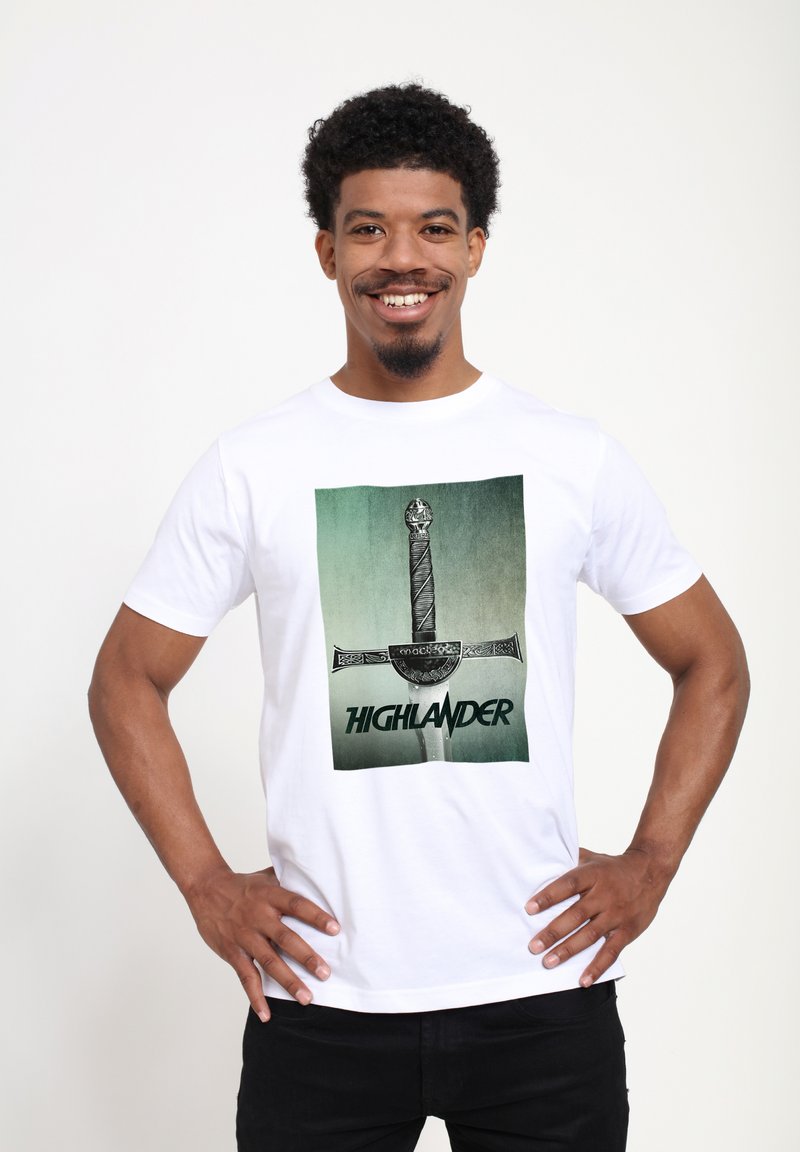 Hvid bomuld T-shirt med en grafisk print af et sværd med teksten "HIGHLANDER" nedenfor. Designet har en gradient grøn baggrund.