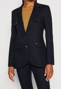 Tmavě modrý přizpůsobený blejzr vyrobený z texturované tkaniny, který má dvoubottoní zapínání, přední kapsy a kovové akcenty.