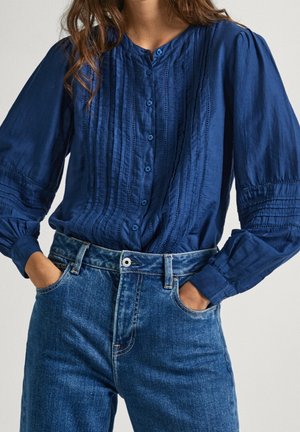 Marineblauwe, gestructureerde blouse met knopen aan de voorkant en verticale plooien, gecombineerd met een hooggetailleerde, lichtblauwe spijkerbroek.