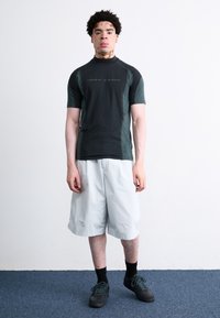 Camisa atlética de manga curta, preta e verde-escuro, com gola alta, combinada com calções cinzentos claros e ténis pretos, contra um fundo simples.