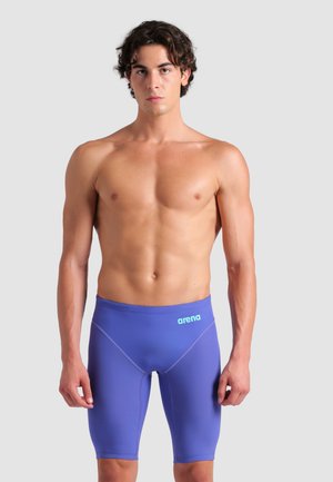 Młody mężczyzna stojący przodem, ubrany w niebieskie kąpielówki typu jammer marki Arena, na tle jednolitego jasnoszarego tła.