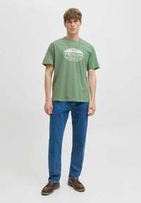 Žalias medvilninis marškinėliai su užrašu "PREMIUM", trumpomis rankovėmis, laisvas aptemdymas, derinami su mėlynomis džinsinėmis kelnėmis ir rudomis bateliais. Paprastas dizainas, kasdienis stilius.