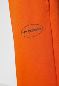 Orange bomullsjoggers med en tryckt "new balance"-logotyp i en grå oval design på vänster sida, vilket betonar en mjuk textur och livfull färg.