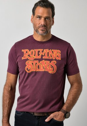 Hombre con barba que lleva una camiseta burdeos de los Rolling Stones y vaqueros oscuros, de pie con una mano en el bolsillo frente a un fondo liso.