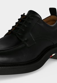 Chaussure habillée noire en cuir à lacets avec détails de couture et un petit accent rouge, blanc et bleu sur la semelle près du talon.