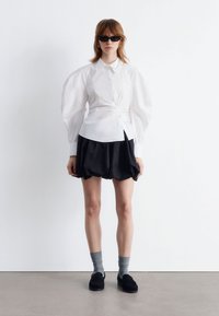 White button-up shirt with puffed sleeves, paired with a black skirt. Model wears grey socks and black shoes, with dark sunglasses.