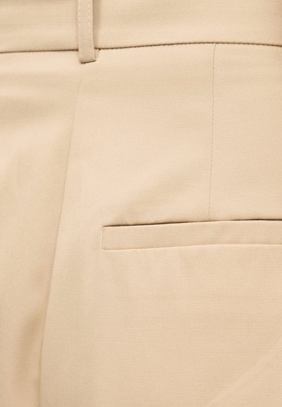 Close-up of beige pants featuring a waistband with belt loop and a horizontal back welt pocket.
