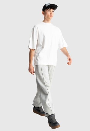 Młoda osoba idąca, ubrana w białą luźną koszulkę, szare spodnie dresowe, czapkę z daszkiem i czarne trampki z brązowymi podeszwami na jednolitym tle.