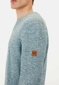 Light blue knitted sweater with a textured pattern, featuring a brown leather logo patch on the left sleeve. Fitted design, crew neckline.