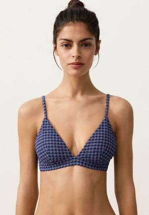 Kobieta o ciemnych włosach związanych w kok, ubrana w niebiesko-fioletowy kraciasty bralet z cienkimi ramiączkami, na jednolitym tle.