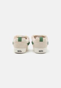 Beige suede sneakers with green accents, featuring a padded collar and rubber sole. Back view shows branding logo on the heel.