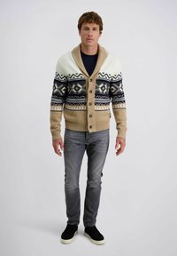 Beige en crème gebreide cardigan met een geometrisch patroon, knoopsluiting en sjawlboord, gecombineerd met grijze jeans en zwarte sneakers.