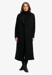 Lange zwarte wollen jas met dubbelgeknoopte voorkant, grote revers en twee zijzakken. Draagt over een zwarte coltrui en broek.